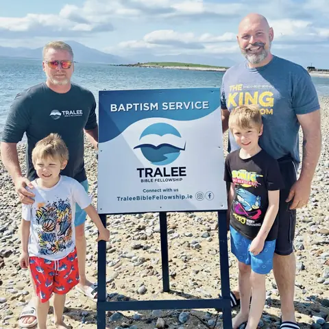 Two men and two boys beside a Baptism Service sign on the beach